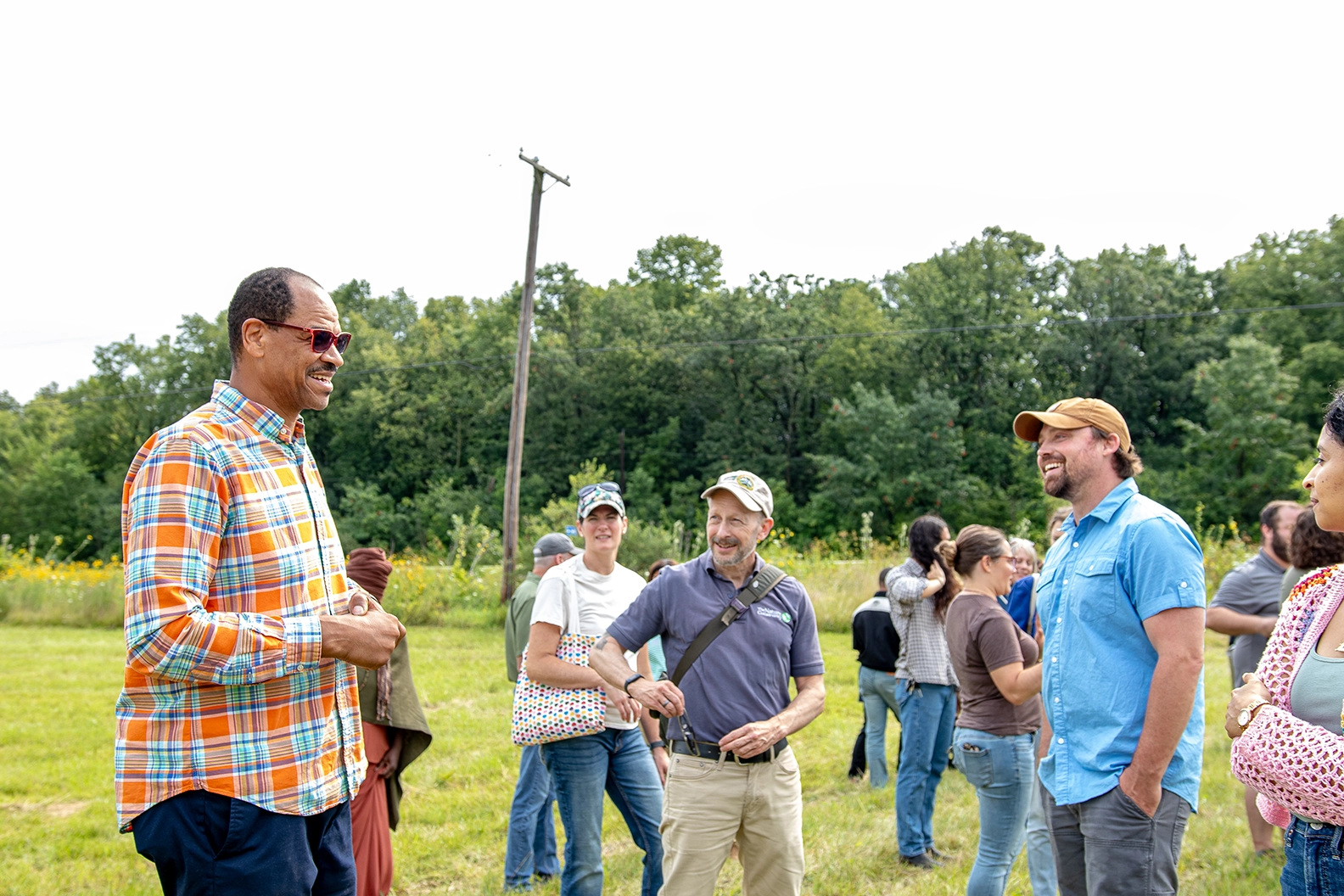 A diverse group of people engaged in conversation outdoors, gathered in a green field surrounded by trees. One man in a colorful plaid shirt is smiling and interacting with others, creating a lively community atmosphere.